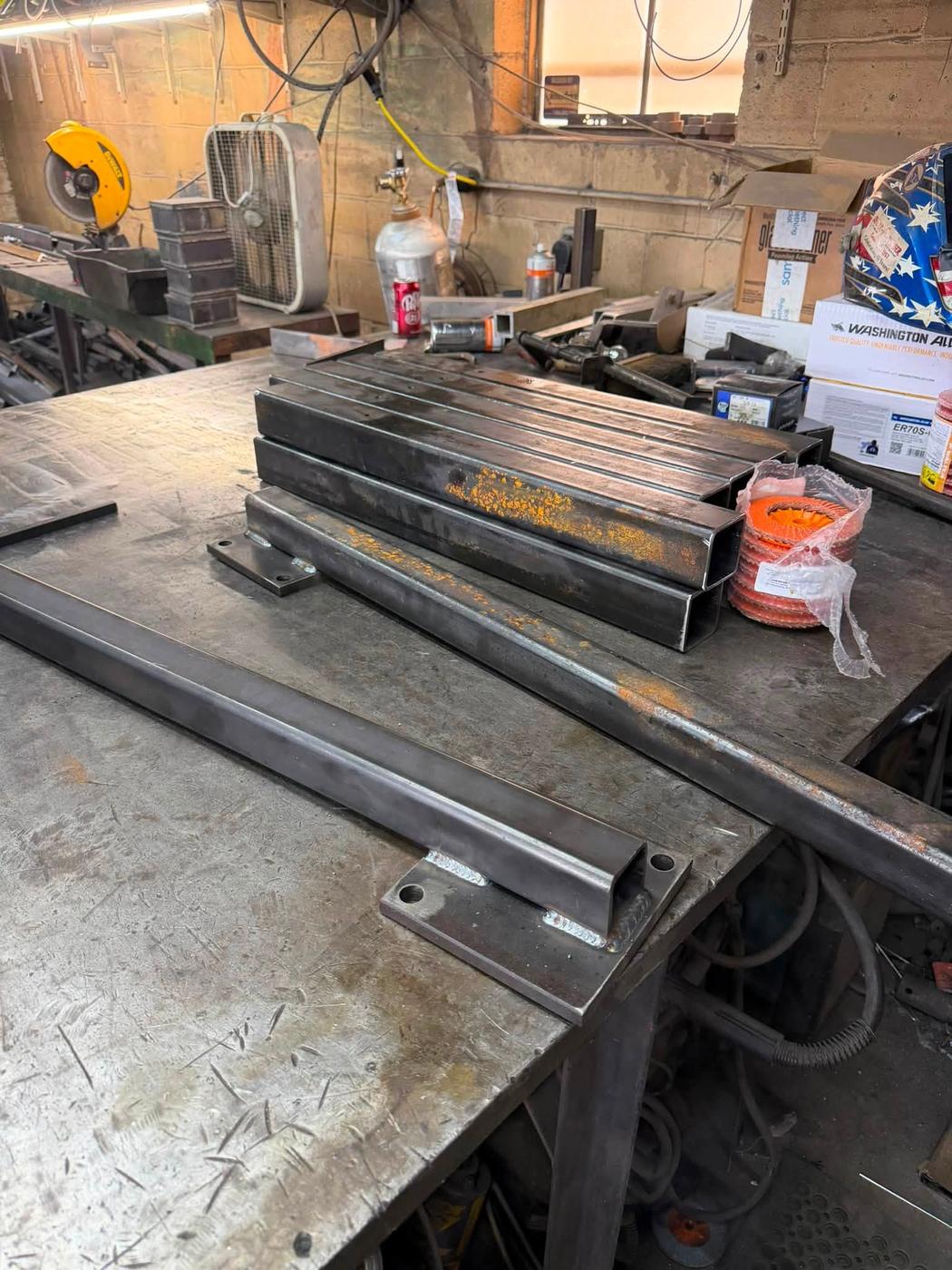 Inside the shop: a steel welding table with fabricated tube frames, a chop saw and welding gear